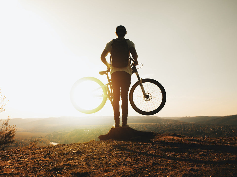 Rear view of a biker holding up his bike while looking into the sun from a hilltop