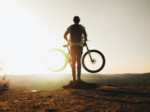 Rear view of a biker holding up his bike while looking into the sun from a hilltop