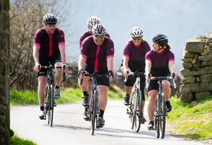 A group of cyclists in matching maroon jerseys smiling and having a conversation riding along a country lane