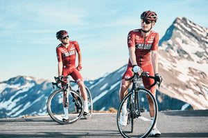 Two male cyclists stopped on a mountain road with snow-peaked mountains in the background.