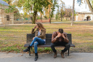 Man and woman arguing on a park bench while the man looks down with has hands on his head and the woman looks away