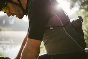 Cyclist with sunglasses on and a camera strapped around his back looking down while he rides his bike