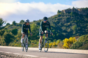 Two cyclists riding and talking happily on a road amid hills